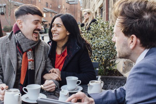 Happy couple are sitting with a financial advisor at a cafe. He is showing them a digital tablet with information and they are laughing while drinking tea.
