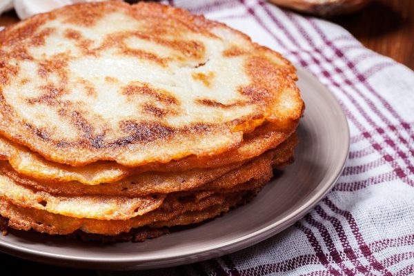 Stack of potato pancakes on a wooden table. In the background potatoes, onions and garlic