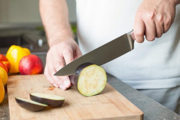 A professional chef quickly cuts eggplants on a wooden board using a ceramic knife. Close-up.