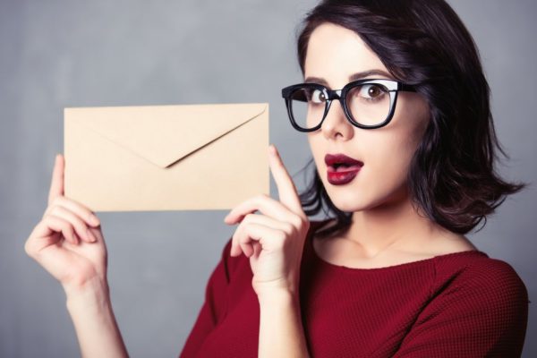 Portrait of young woman in black dress with envelope Christmas gift, surprised over grey background
