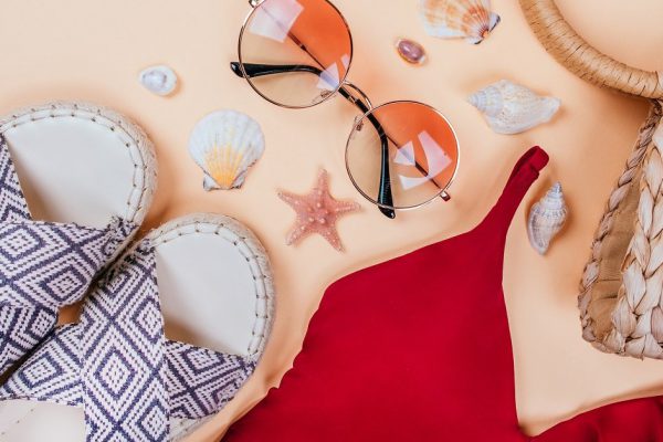 Summer fashion flatlay with gradient round sunglasses, straw bag with pompoms and red swimsuit with frills on beige background. Decorated with sea shells. Perfect beach set for holidays. Marina style.