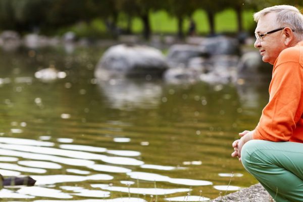 Profile of aged man in glasses sitting near pond in park watching ducks