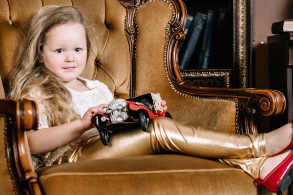 Little baby girl sitting on retro golden chair and playing with red car vintage toy alone