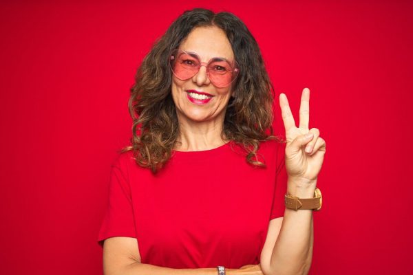 Middle age senior woman wearing cute heart shaped glasses over red isolated background smiling with happy face winking at the camera doing victory sign. Number two.