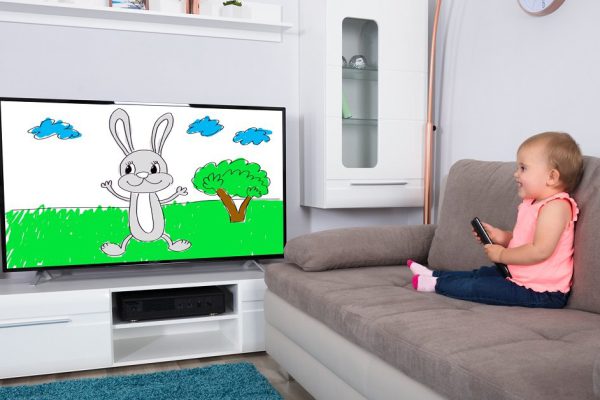 Happy Baby Girl Sitting On Sofa Watching Cartoon On Television