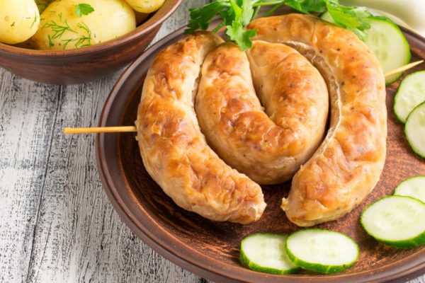 Young boiled potatoes with dill in the bowl, sausage and cucumber on the plate, parsley and onions on the wooden board