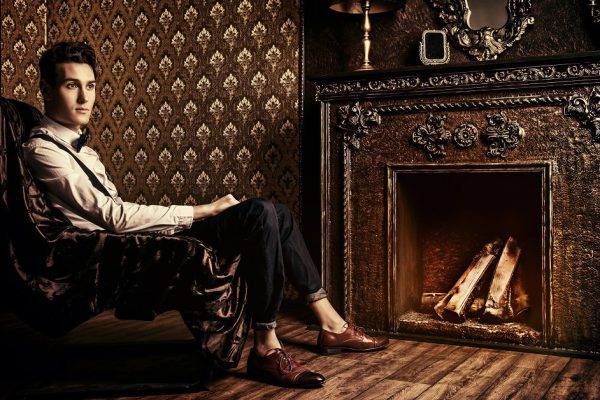 Elegant handsome young man sitting by the fireplace in a room with classic vintage interior. Fashion shot.