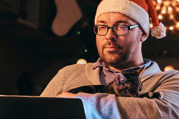 Man in Santa hat and glasses dressed in warm sweater hugging cat and using laptop celebrating Christmas Eve at home.