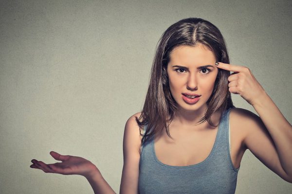 Closeup portrait of angry mad young woman gesturing with her finger against temple asking are you crazy? Isolated on gray wall background. Negative emotions facial expression feeling body language