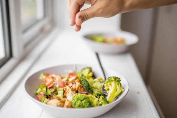 Hand gently adding a pinch of salt to broccoli and chicken salad dish next to window. Nutritious dish with vegetables and meat on white plate. Balanced diet