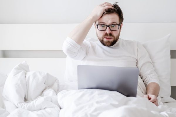 Upset man with a laptop in bed received bad news, he is confused, holding hand to his head and watching the screen in shocked eyes.