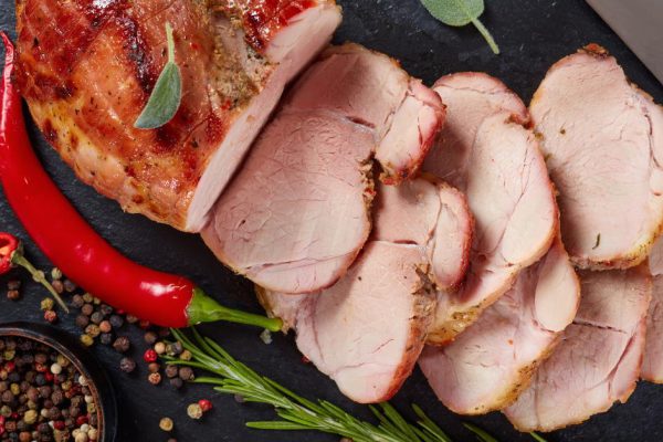 delicious juicy roasted piece of pork meat cut in slices on a black slate cutting board with knife, chili pepper, peppercorns, fresh herbs, view from above, close-up