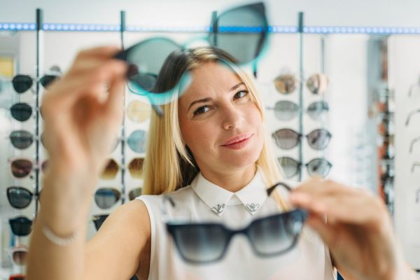 Female buyer chooses sunglasses in optics store, showcase with spectacles on background. Selection of eyes protection in glasses shop, optometry concept
