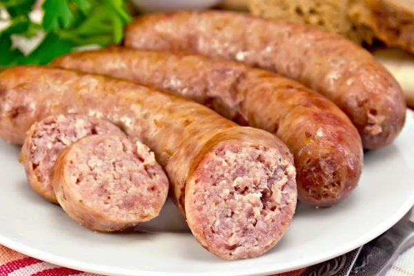 Pork sausages fried in a dish, bread, sauce, tomato, parsley, fork, knife, napkin on the background of wooden boards