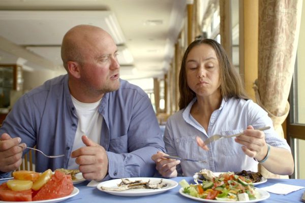 The husband bald and in glasses and the dark-haired middle-aged wife, sit in cafe, eat, treat each other, estimate food, laugh, communicate. Have them on the table are worth fruits, vegetables and hotter.