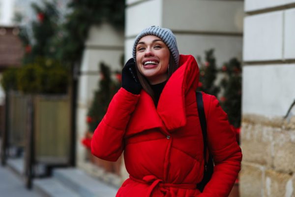 Portrait of laughing woman on background of Christmas Fair talking on mobile phone. Woman in red winter jacket standing on street on winter day. Christmas holidays concept. Copy space on left side