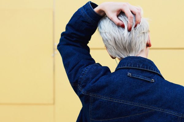 Rear view of female blonde hipster dressed in trendy jeans jacket with copy space area for your brand name or advertising posing on urban setting wall background. Female with stylish haircut posing