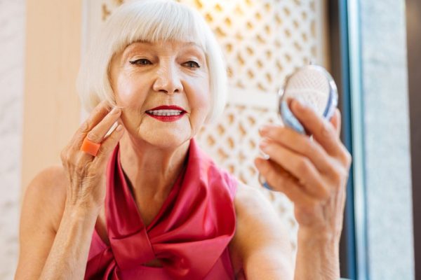 My face. Joyful elderly woman touching her cheek while looking at her face in the mirror