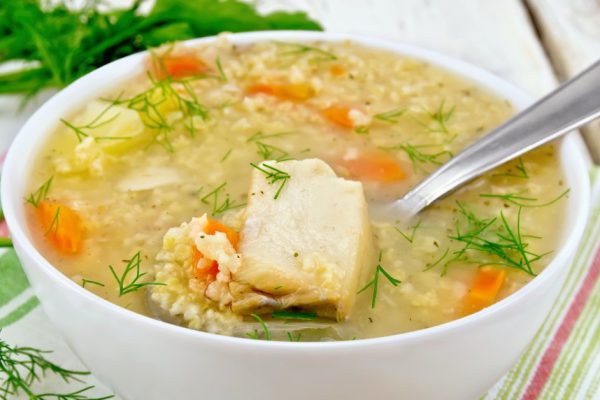 Soup fish kulesh with millet, potatoes and carrots and spoon in a bowl on a napkin, parsley, dill on a wooden board background