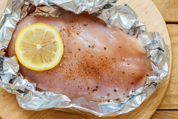 Raw chicken fillet with lemon slice, black pepper and salt in baking foil. Uncooked chicken breasts on wooden cutting board