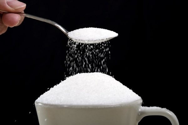 man hand with spoon pouring a crazy lot of it spilling out everywhere in full coffee cup in insane sugar addiction and unhealthy nutrition concept isolated on black background