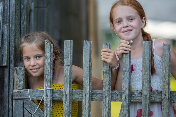 Two little cute girlfriends look out for wooden village fence.