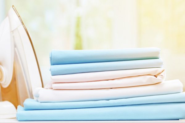 Close-up of blue and white clean bedding on a blurred background. A stack of folded bedsheets on the table, an iron stands nearby. Sunlight from the window.