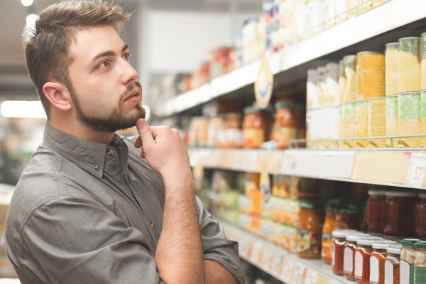 Buyer man looks at the supermarket shelf and thinks what product to take. Buyer selects canned food at the store. Shopping in a supermarket.