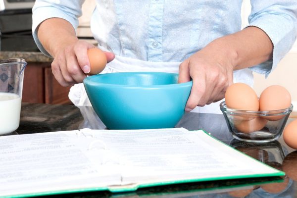 A homemaker cracks an egg into a mixing bowl while following the instructions of a cookbook.