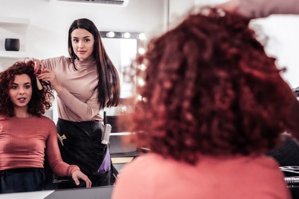 Professional hair styling. Joyful positive woman looking at her reflection in the mirror while visiting a hair stylist
