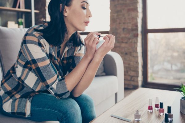 Profile photo of cute lady made correction finger nails applying red color, polish blowing helping dry wear casual clothes sitting cozy sofa apartment indoors
