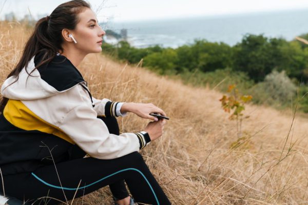 Photo of nice caucasian woman in sportswear using earpods and smartphone while sitting on dry grass at seashore