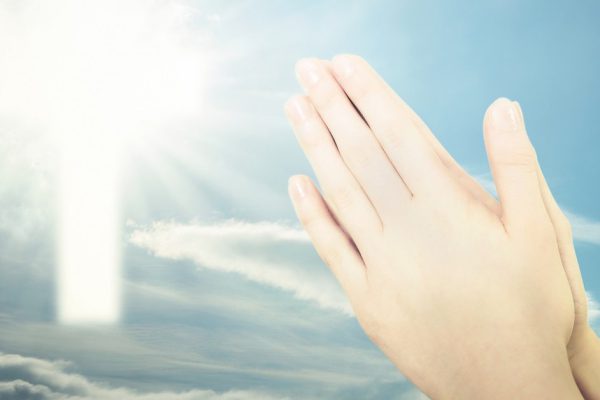 Concept of Christian prayer - folded hands for prayer against a blue sky and a shining cross sign (mixed)