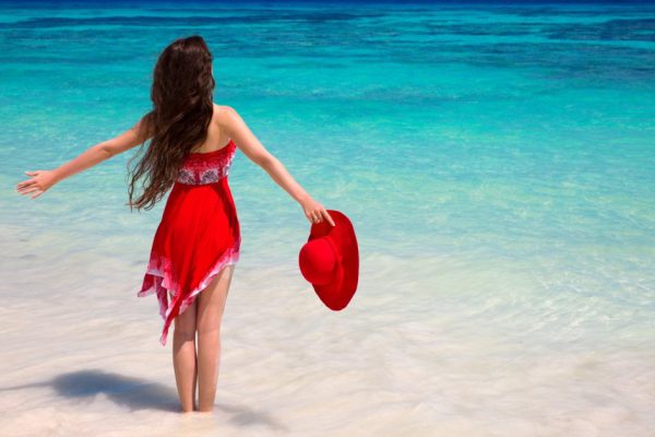 Happy woman enjoying on exotic beach in summer by tropical blue water. Attractive girl in red dress resting, outdoor portrait. Bliss freedom concept. Travel.