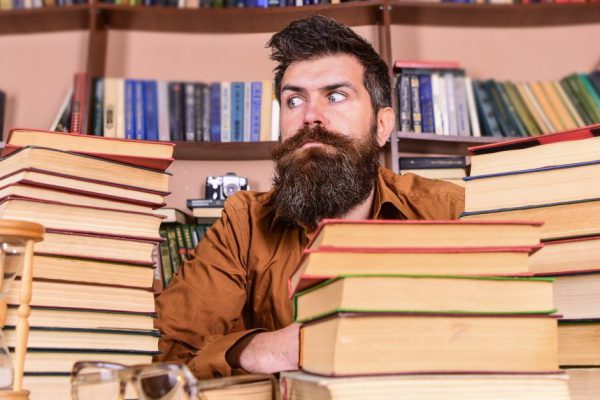 Bookworm concept. Man on strict face sit between piles of books, while studying in library, bookshelves on background. Teacher or student with beard sit at table with hourglass and glasses, defocused.