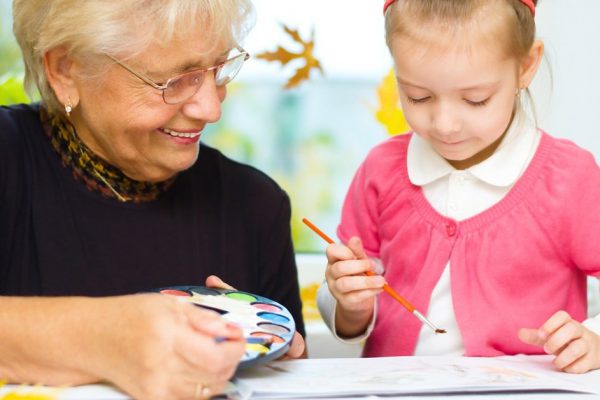 Grandmother with granddaughter painting with paintbrush and colorful paints, autumn background