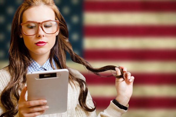 Picture of pretty girl in big hipster glasses with ebook. Young student reading on USA flag blurred background.