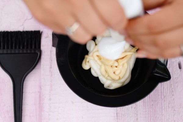 Picture of woman's hands mixing dyes for hair in black bowl. Closeup of hairdresser's equipment on pink plank table background.