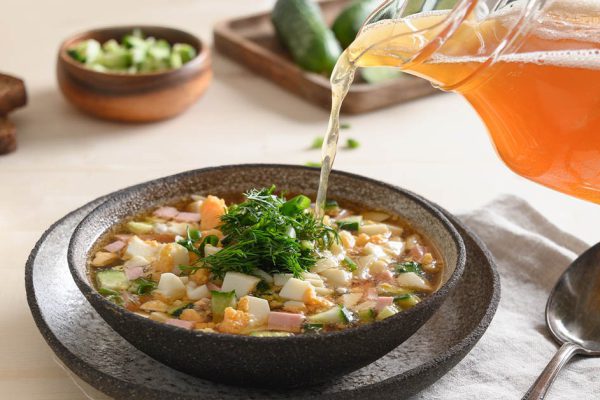 Traditional Russian cold soup okroshka. Kvass in the process of pouring in the plate. Sour cream, horseradish, salt, bread and herbs on background