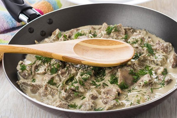 Chicken liver in sour cream, spoon in frying pan, empty plate on wooden table