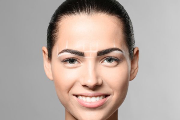 Young woman before procedure of eyebrows permanent makeup on grey background