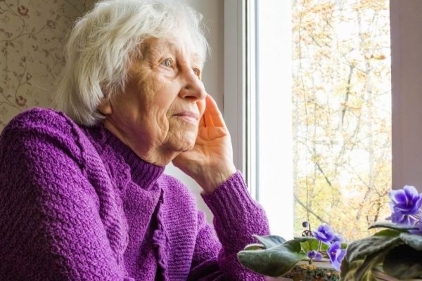 Old lonely woman sitting near the window in his house with flowers