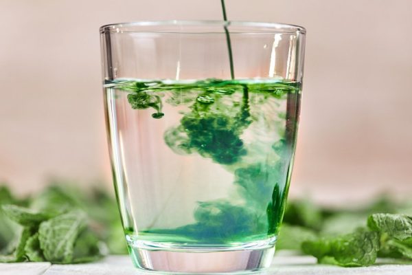 Dissolving liquid chlorophyll in glass of water on white table with green mint