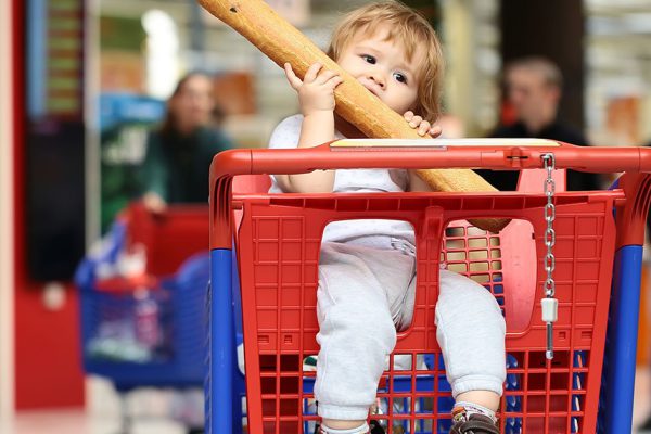 Closeup beautiful cute baby holding and biting French bread sitting in red and blue shopping trolley against supermarket full of people background, horizontal picture