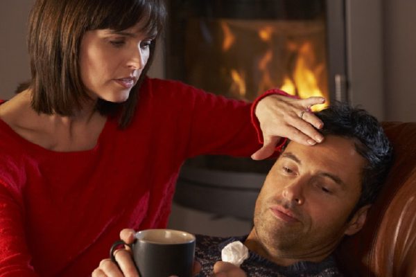 Wife Nursing Sick Husband With Cold Resting On Sofa By Cosy Log Fire