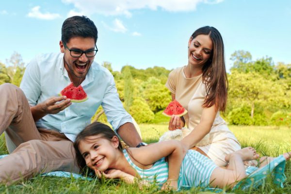 Family In Park. Happy Beautiful Young Parents And Smiling Child Having Fun On Picnic In Park, Kid Eating Fruits. People Relaxing And Spending Time Together Outdoors, In Nature. Relationship Concept