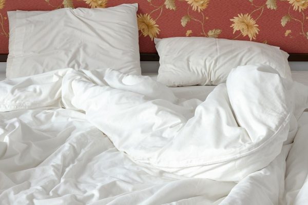 Unmade / untidy bed with a white crumpled blanket and two messy pillows in a bed room. Bedclothes are not neatly arranged for new customers / guests to sleep in. View from the foot of the bed.