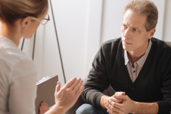 Discussing problems. Handsome troubled upset man sitting on the sofa and holding his hands together while listening to his psychologist