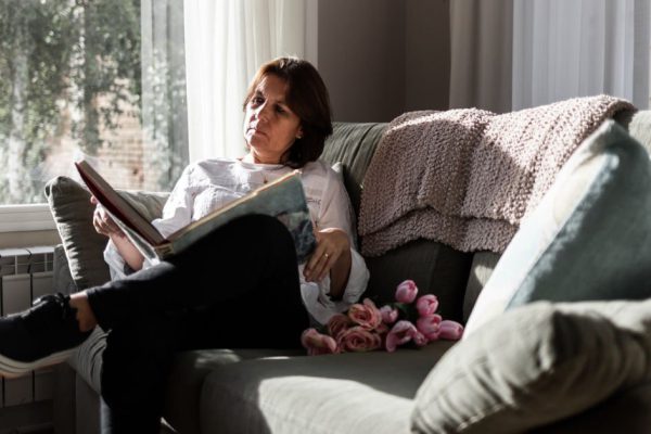 Light-eyed, brown-haired woman in a white shirt reading a book comfortably from her home couch. She complies with the social isolation.