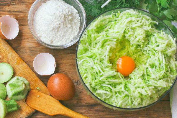 Cooking homemade pancakes from courgettes. Grated zucchini in a bowl, eggs, herbs, flour on a wooden table. Top view